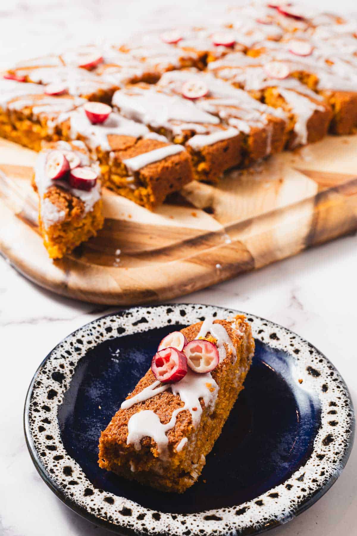 A slice of pumpkin spice cake topped with white icing and sliced red fruit is placed on a black and white speckled plate. In the background, more slices of the gluten-free cake with similar toppings are arranged on a wooden board.