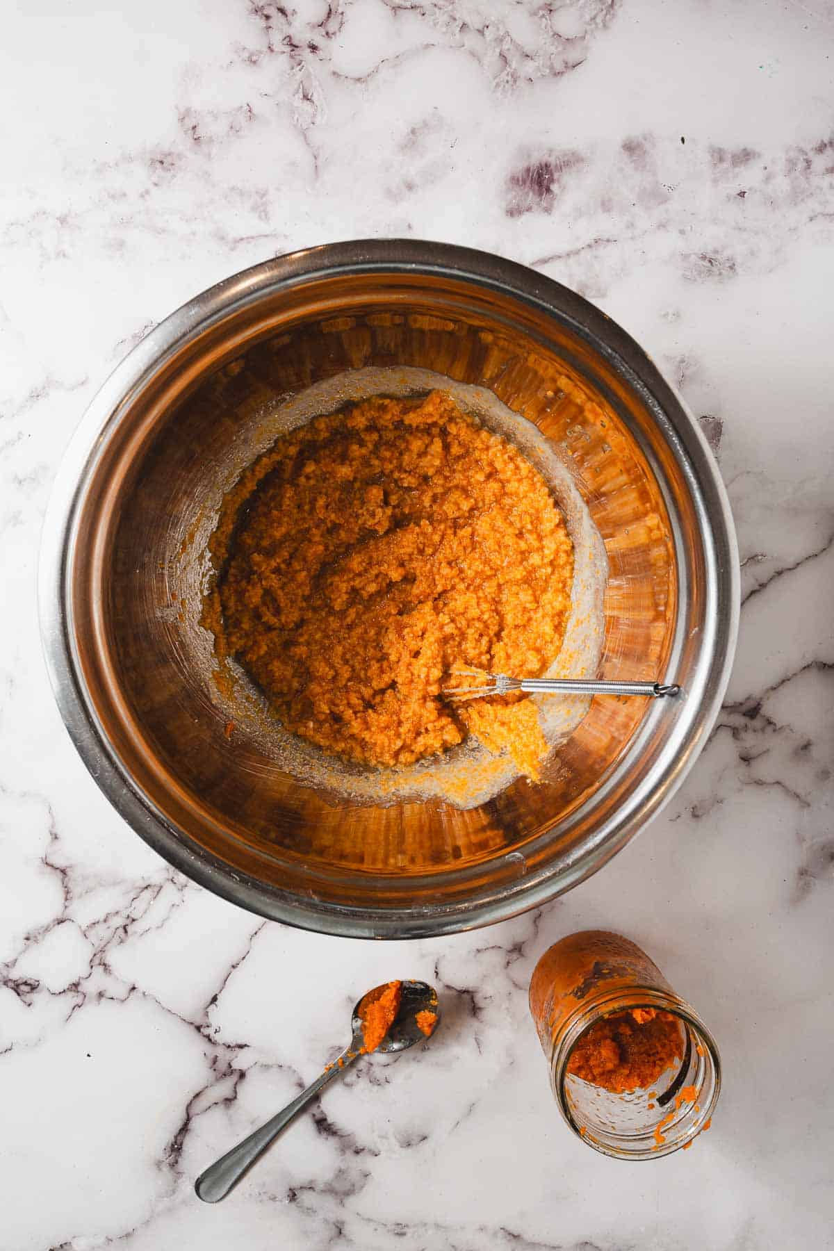 A metal bowl filled with gluten-free orange-red chili paste, accompanied by a silver spoon, sits on a marble countertop. Next to the bowl, a small jar filled with the same paste and a spoon with some paste on it are placed nearby.