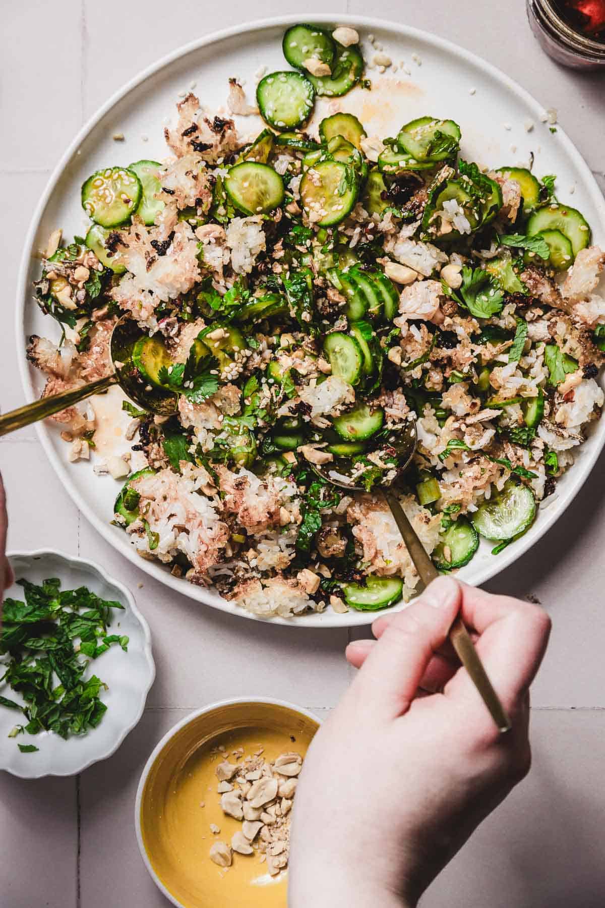 A person's hand holding a fork is poised to enjoy a crispy cucumber rice salad, featuring sliced cucumbers, rice, and herbs. Nearby, a small bowl of chopped greens and a sprinkling of peanuts complement the dish on the light surface.
