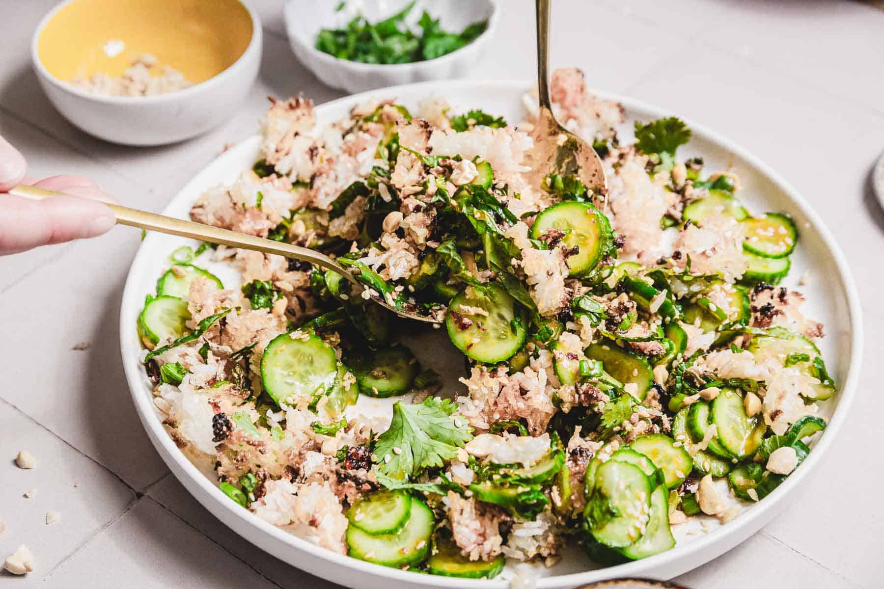 A white plate features a crispy cucumber rice salad served with sliced cucumbers, cilantro, and peanuts. Utensils rest beside it, while small bowls of chopped herbs and peanuts decorate the neutral-toned surface.
