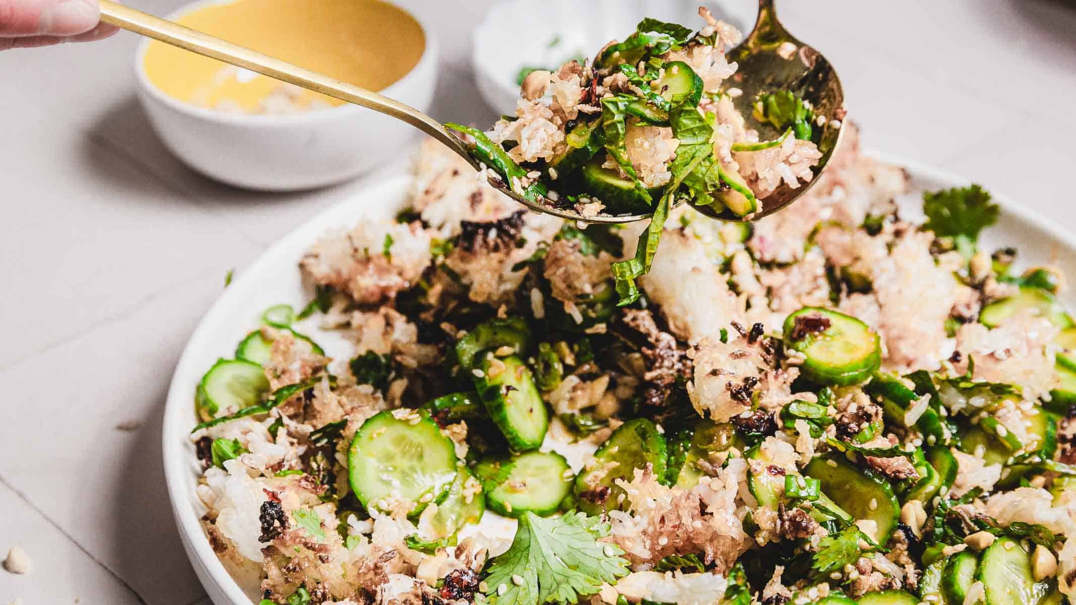 A serving spoon lifts a portion of the Crispy Cucumber Rice Salad recipe, showcasing sliced cucumbers, rice, herbs, and crunchy elements from a white plate. In the background, a small white bowl with a yellow interior complements the vibrant dish.