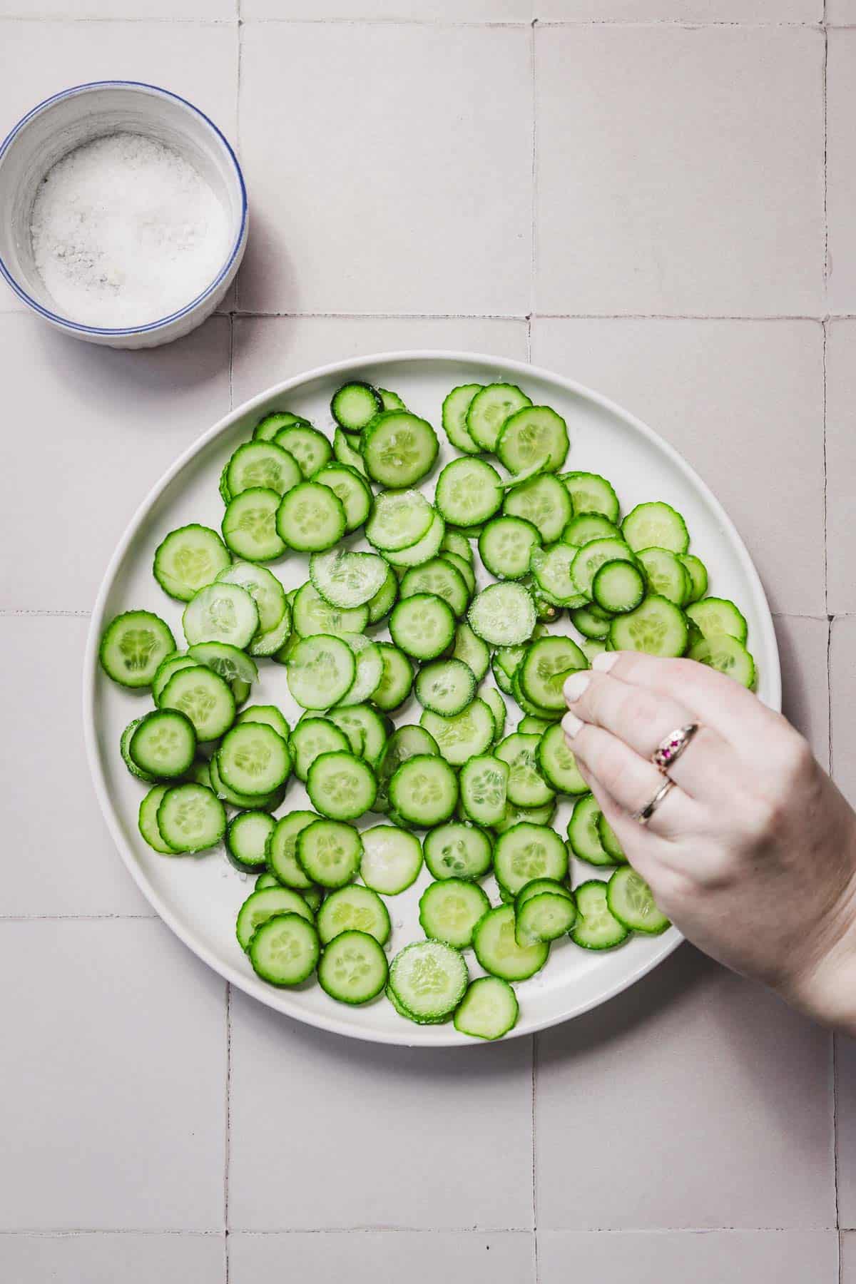 A hand sprinkles salt onto a plate of sliced cucumbers, ready for the deliciously refreshing Crispy Cucumber Rice Salad. The plate rests on a tiled surface, with a small bowl of salt nearby.