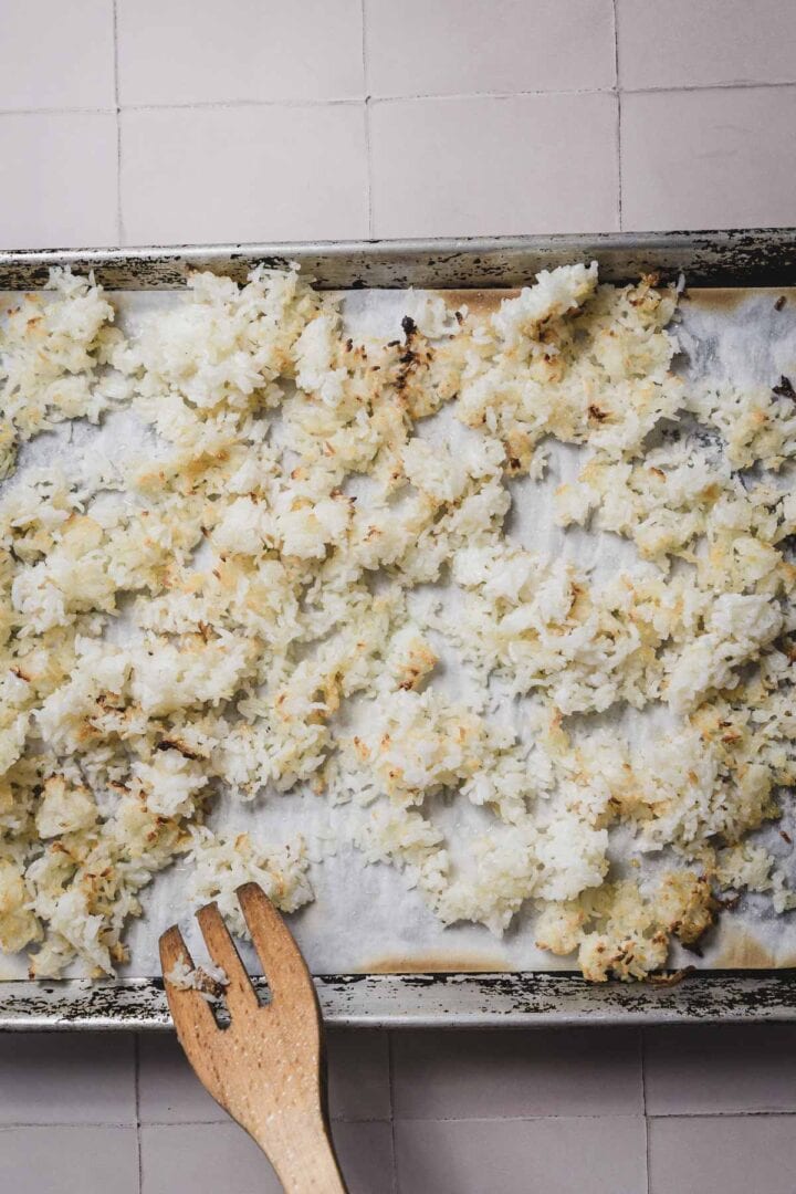 Baked shredded coconut is spread on a parchment-lined baking sheet, ready to add crunch to your Crispy Cucumber Rice Salad Recipe. A wooden spatula rests on the corner of the sheet. The white and lightly browned coconut has a perfectly crispy texture against the tiled surface backdrop.