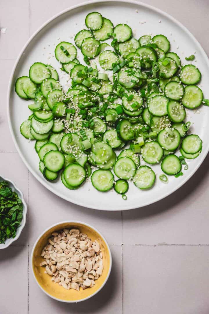 A white plate displays a crispy cucumber rice salad, artfully topped with sesame seeds and chopped green onions. Nearby, small bowls hold chopped peanuts and herbs, creating a harmonious arrangement on a light-colored surface.