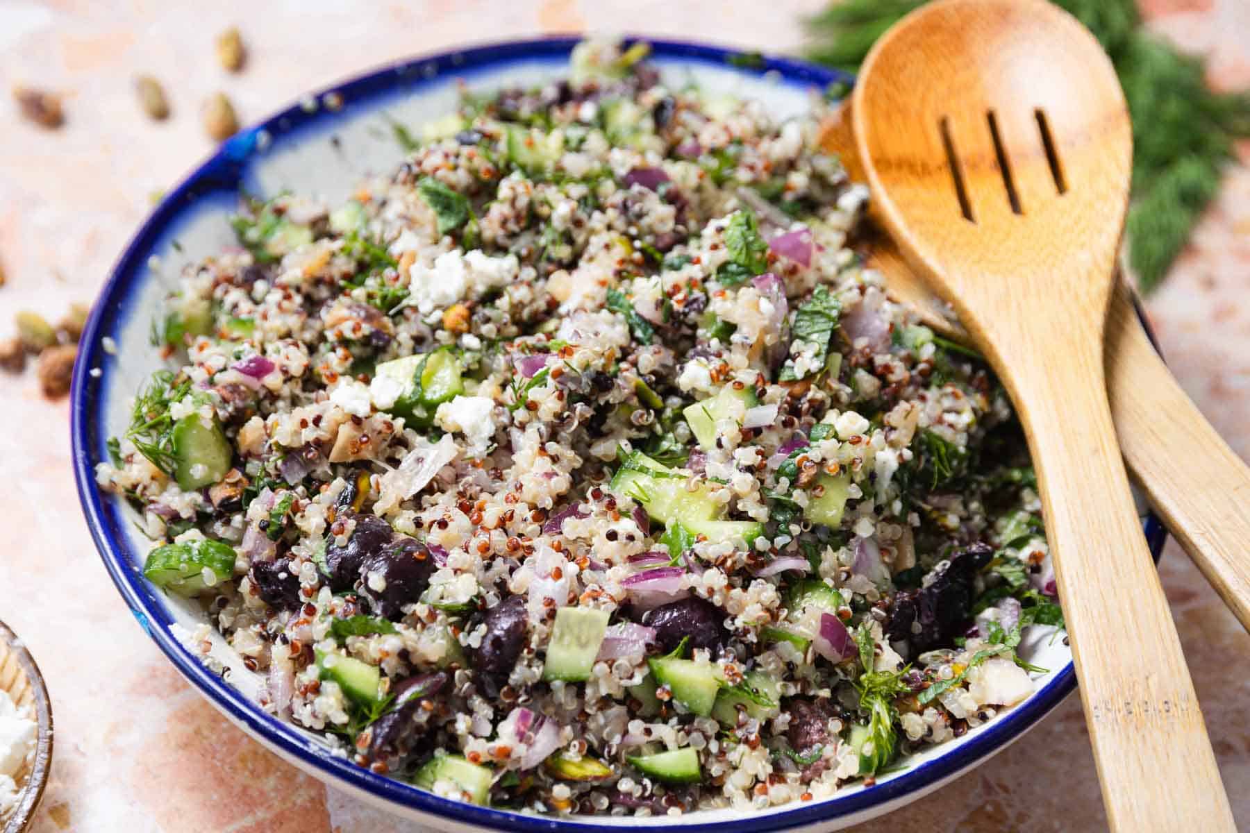A vibrant quinoa cucumber salad brimming with chopped cucumbers, red onions, black olives, and herbs sits invitingly on the table. Two wooden serving spoons rest beside the bowl, complementing the salad's fresh and colorful appearance.