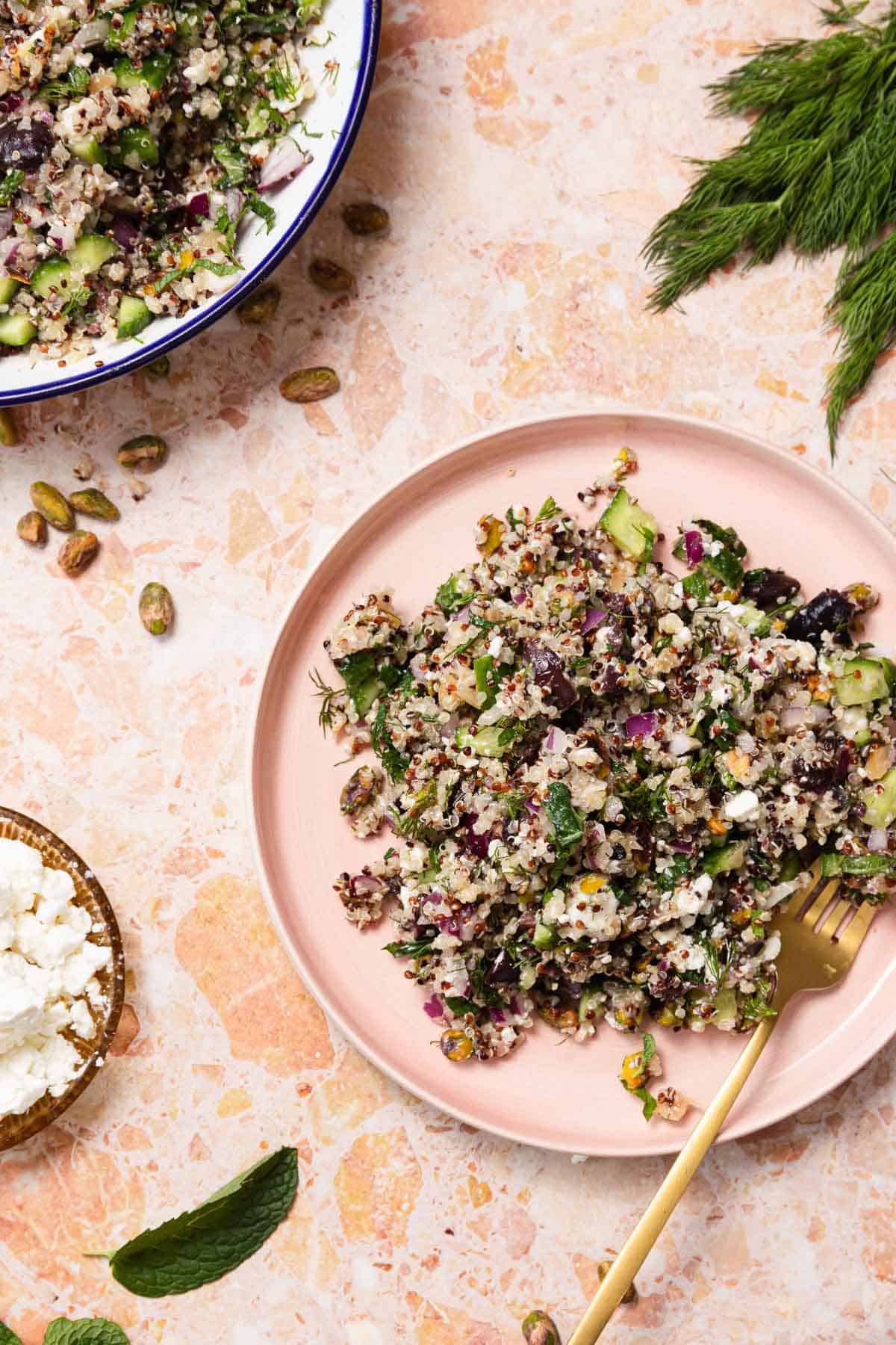 A plate featuring a vibrant quinoa cucumber salad with red onion, herbs, and pistachios rests on a marble surface. A fork lies beside it, ready to dive in. Nearby, a bowl of crumbled feta cheese and sprigs of dill complete this refreshing dish.