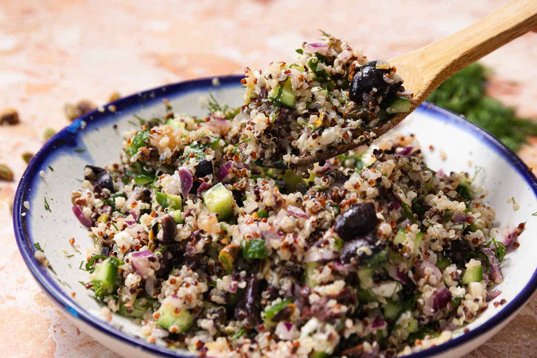 A bowl of quinoa cucumber salad with red onions, black olives, and fresh herbs. A wooden spoon lifts a portion of the vibrant mix. The background features a light pink and orange surface, enhancing the dish's colorful appeal.