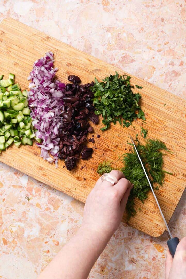 A person is chopping fresh dill on a wooden cutting board, preparing for a vibrant quinoa cucumber salad. Next to the dill are piles of chopped cucumber, red onion, black olives, and parsley. The board rests elegantly on a marbled countertop.
