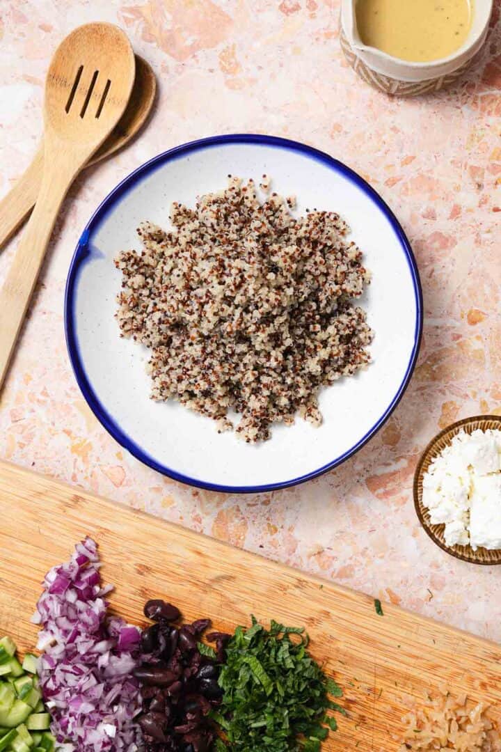 A bowl of cooked quinoa is placed on a stone countertop, perfect for a refreshing Quinoa Cucumber Salad. Nearby are wooden utensils, chopped red onions, green herbs, sliced black olives, diced cucumber, a small bowl of feta cheese, and a bowl of dressing.