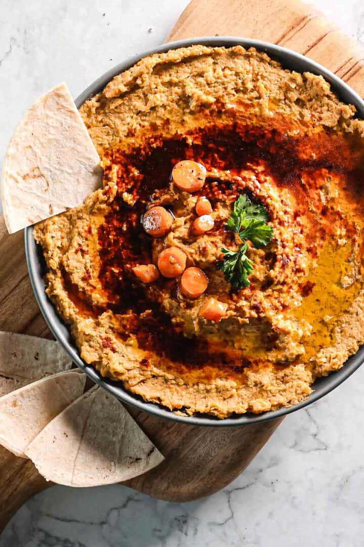 A bowl of hummus garnished with chickpeas, parsley, and paprika sits on a wooden board alongside roasted carrots. Several pieces of pita bread are placed nearby. The hummus has a swirl pattern with oil on top, beautifully contrasted against the marble background.