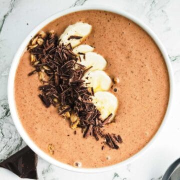 A bowl of chocolate peanut butter smoothie topped with sliced bananas, granola, and grated chocolate rests on a marble surface. A piece of chocolate is nearby on the table.