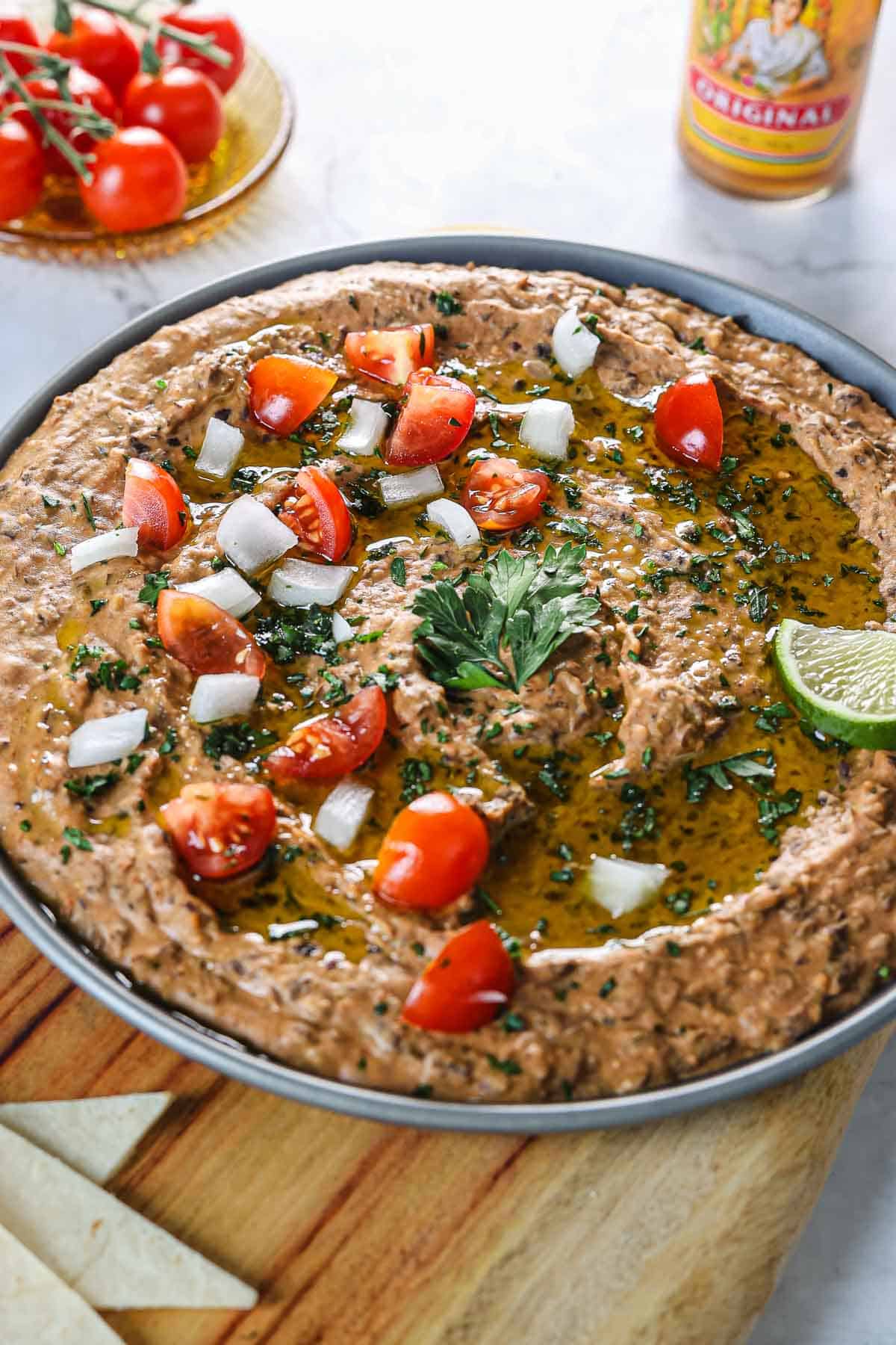 A Mexican-style dish featuring a black bean dip garnished with chopped tomatoes, diced onions, and parsley, drizzled with olive oil. A lime wedge is on the side. Cherry tomatoes are in the background, along with a bottle of hot sauce. Tortilla chips are visible in the corner.