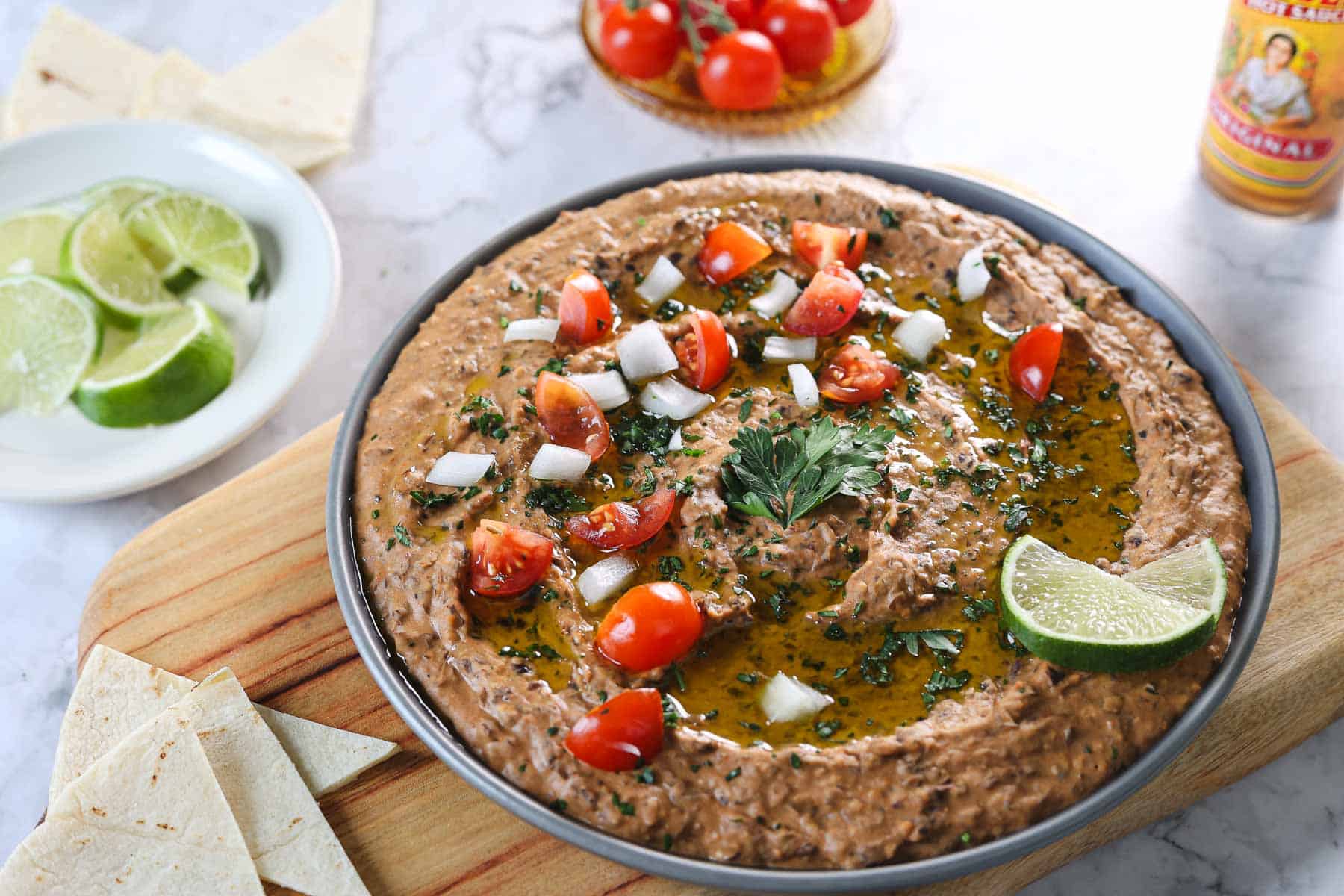 A bowl of Mexican-style refried black bean dip topped with chopped tomatoes, onions, and cilantro is garnished with lime wedges. Placed on a wooden board, it comes with tortilla pieces and lime slices. Cherry tomatoes and hot sauce add extra zest in the background.