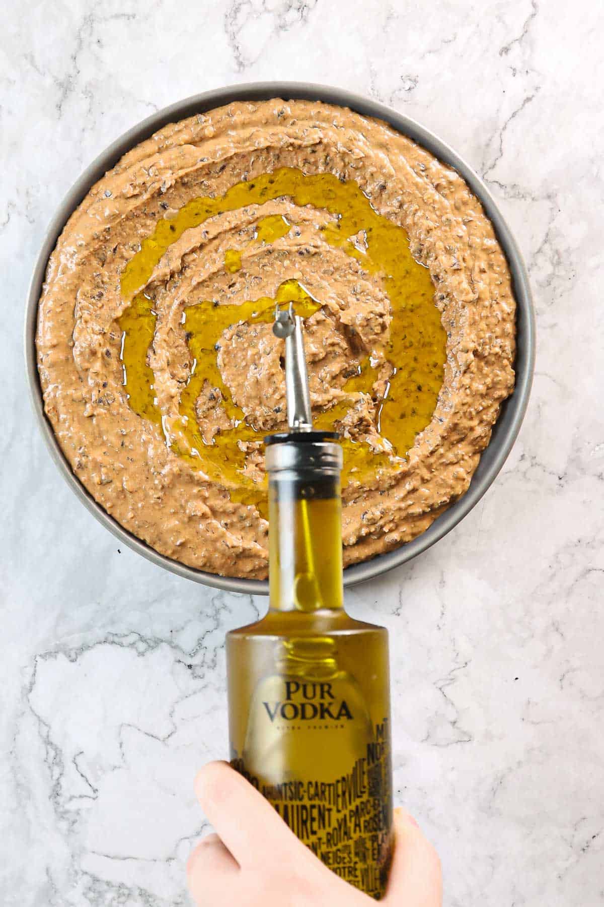 A hand holds a bottle of olive oil over a round baking pan filled with Mexican-style, brown, textured batter resembling black bean hummus. The oil is being poured in a spiral pattern against the light marble background.