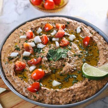A round dish filled with creamy hummus, garnished with chopped tomatoes, onions, and herbs. A slice of lime is on the side. Olive oil is drizzled on top, and cherry tomatoes are visible in the background for a Mexican-style twist.