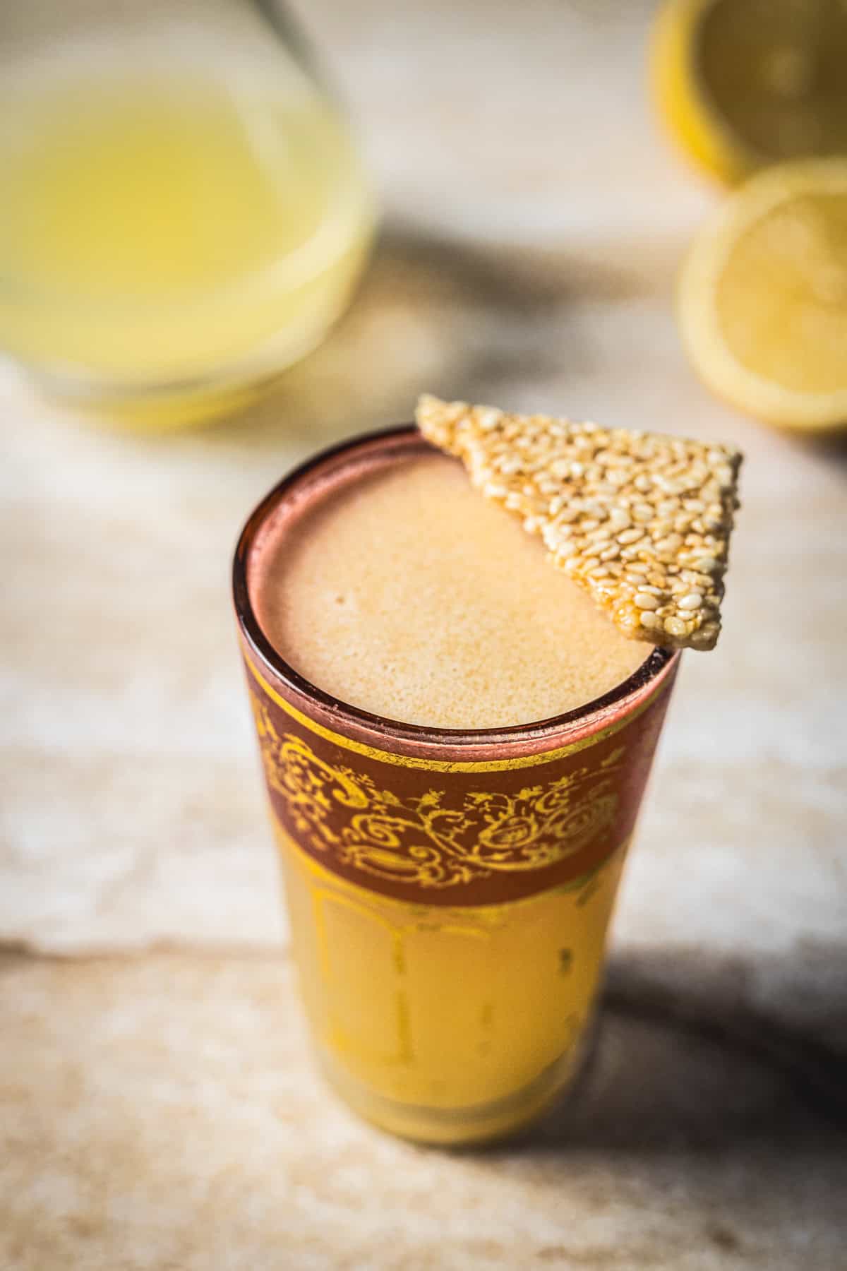 A tall glass filled with a frothy beige 1001 Arabian Nights Cocktail, garnished with a triangular sesame seed brittle, sits on a light surface next to a cut lemon and another cocktail in the blurred background.