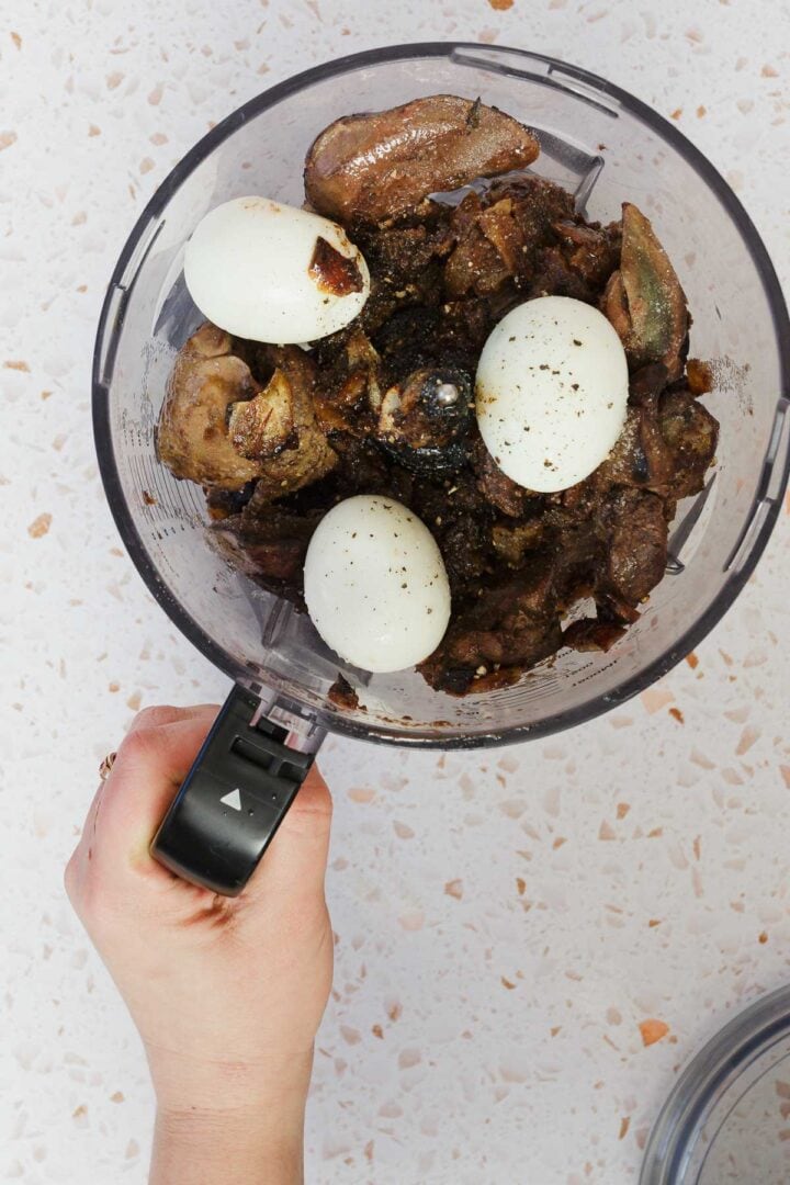 A hand holds a food processor bowl filled with classic Jewish chopped chicken livers, enriched with boiled eggs and onions. The savory blend is perfectly seasoned with ground pepper, set against a speckled white countertop.