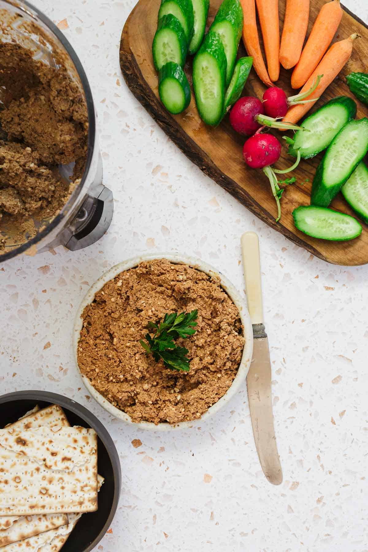 A food processor with Classic Jewish Chopped Chicken Livers spread is next to a bowl of the same topped with parsley. A knife rests nearby. A wooden board displays cucumber slices, carrots, and radishes, while a bowl of crackers is visible below.