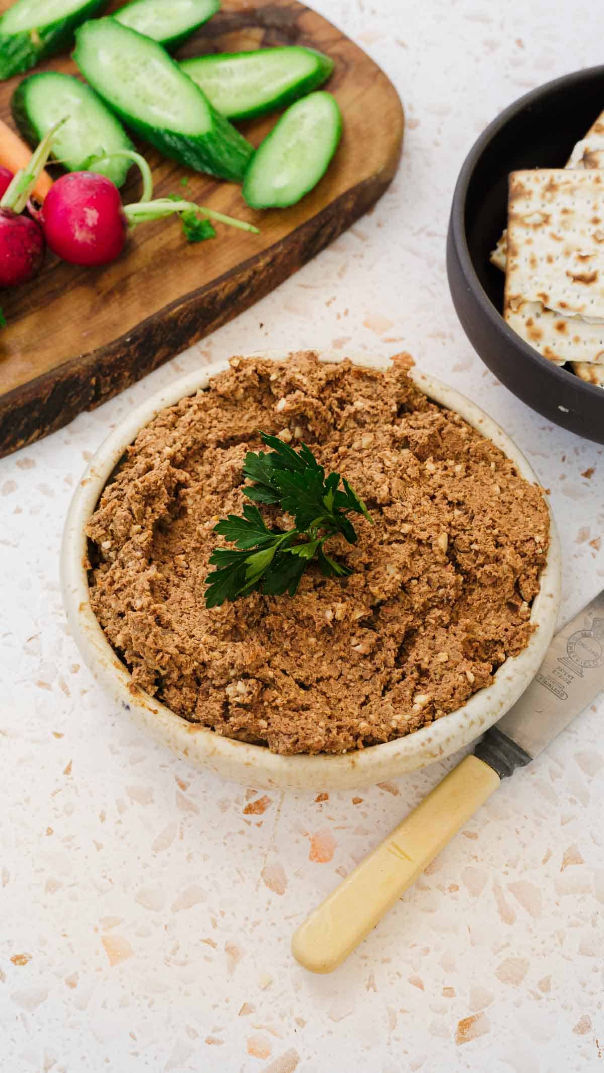 A bowl of classic Jewish chopped chicken livers garnished with parsley sits on a white surface. Nearby, a tray holds sliced cucumbers and radishes, and a plate contains crackers. A knife with a light-colored handle rests beside the bowl.