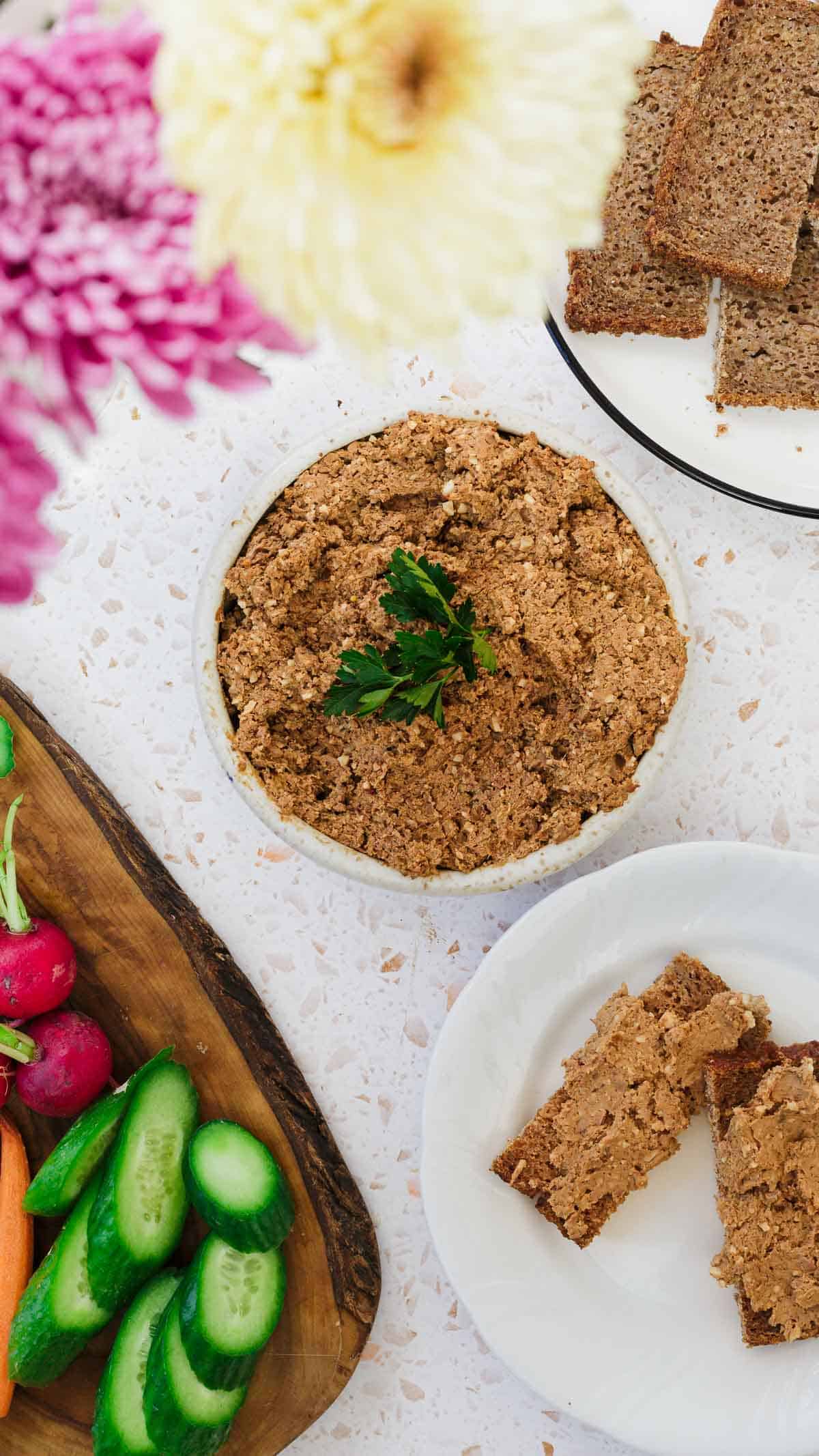 A bowl of classic Jewish chopped chicken livers garnished with parsley sits on a speckled countertop, surrounded by sliced bread, radishes, cucumber slices, and carrots on a wooden board. A flower vase is partially visible at the top left corner.