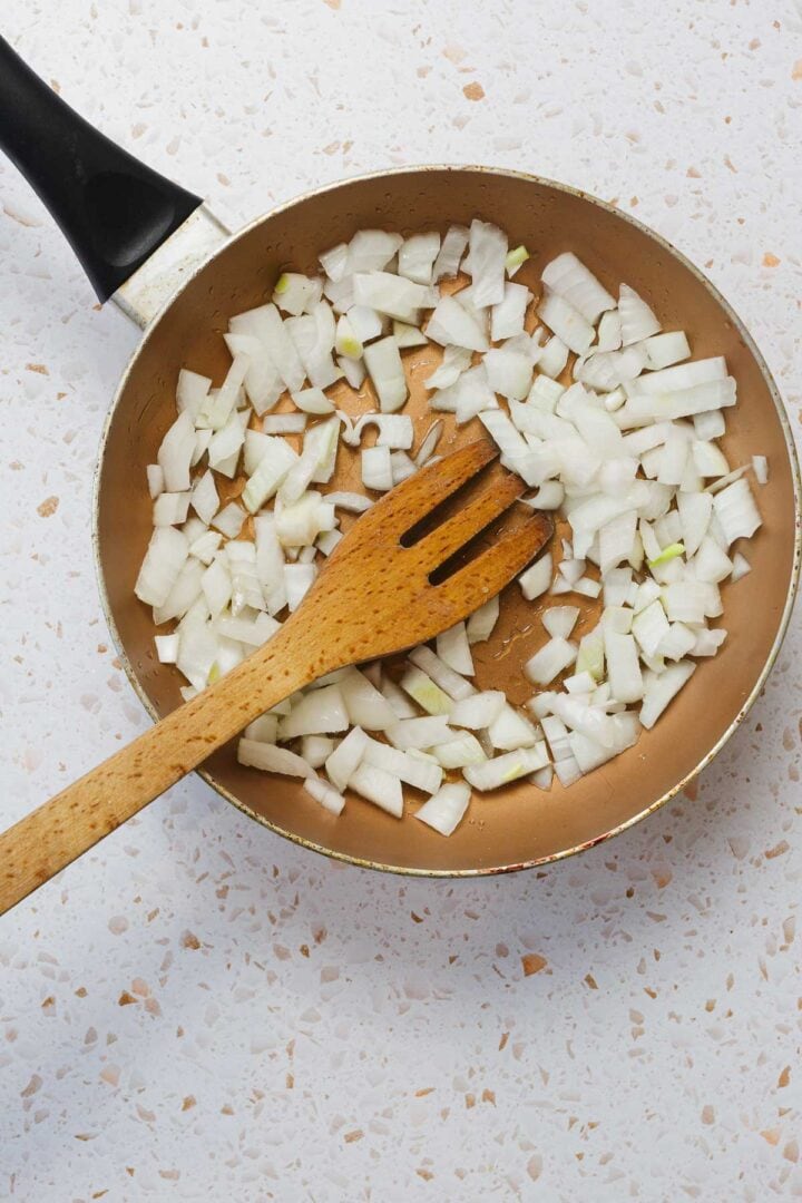 A frying pan on a speckled countertop contains chopped onions, reminiscent of the beginning steps in making Classic Jewish Chopped Chicken Livers. A wooden spatula rests in the pan among the onions, ready to continue this culinary tradition.
