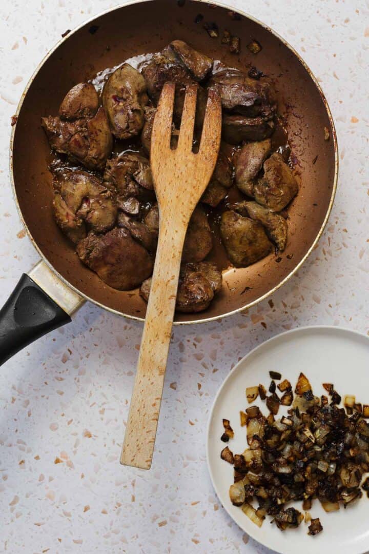 A frying pan with Classic Jewish Chopped Chicken Livers and a wooden spatula rests on a speckled countertop. A small white plate next to the pan holds sautéed chopped onions.