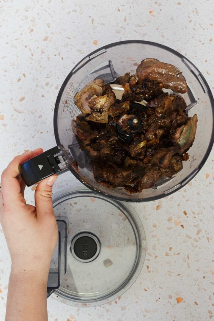 A hand grips the handle of a food processor bowl brimming with classic Jewish chopped chicken livers. The bowl rests on a white speckled countertop, its lid lying beside it.