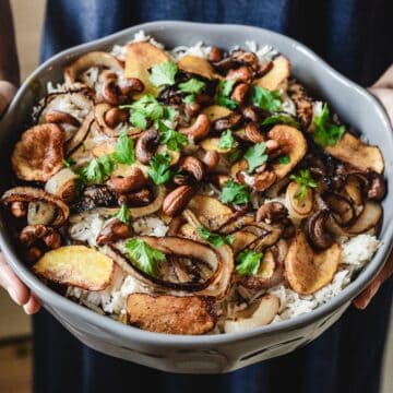 Close-up of a bowl filled with vegetarian delight featuring rice topped with caramelized onions, fried potato slices, roasted mushrooms, cashews, and garnished with parsley. This delectable biryani-inspired dish is cradled lovingly with two hands.