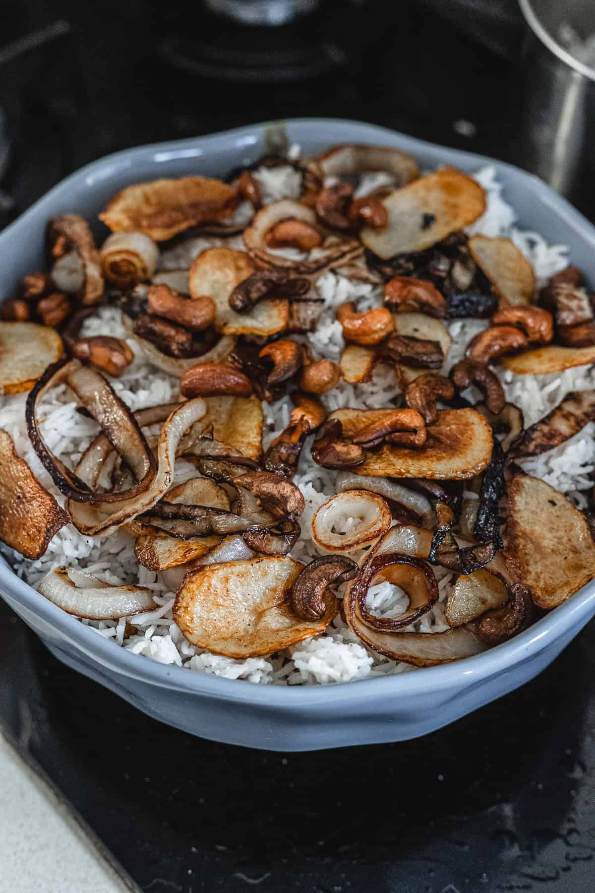 A blue dish filled with fragrant vegetarian biryani, showcasing cooked rice topped with browned onion slices, roasted cashews, and sautéed potato slices. The contents are centered in the dish, and the photo captures it beautifully on a dimly lit stovetop.