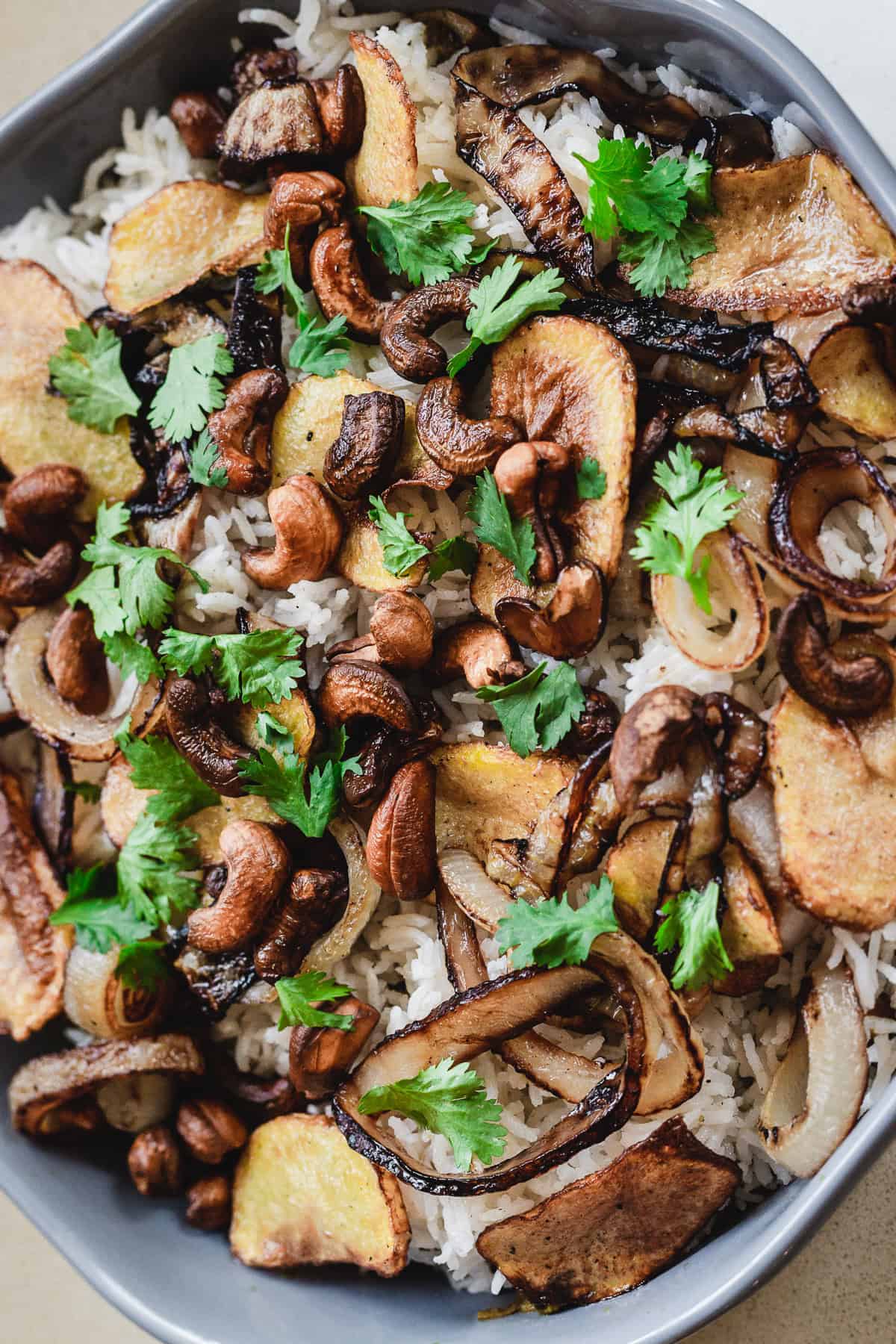A bowl of vegetarian rice topped with fried onions, cashews, sliced potatoes, and fresh cilantro. This biryani-inspired dish is garnished with leafy greens, showcasing a mix of golden-brown and green hues for a visually appealing presentation.