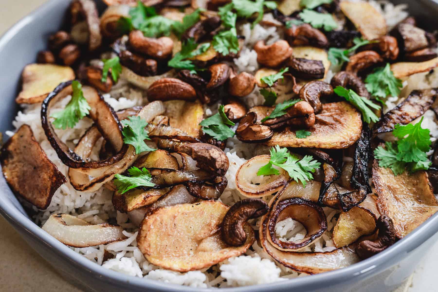 A delightful vegetarian dish featuring white rice layered with sliced, fried onions, earthy mushrooms, and golden-brown potato slices, all garnished with fresh cilantro leaves—offering a hint of biryani flair.