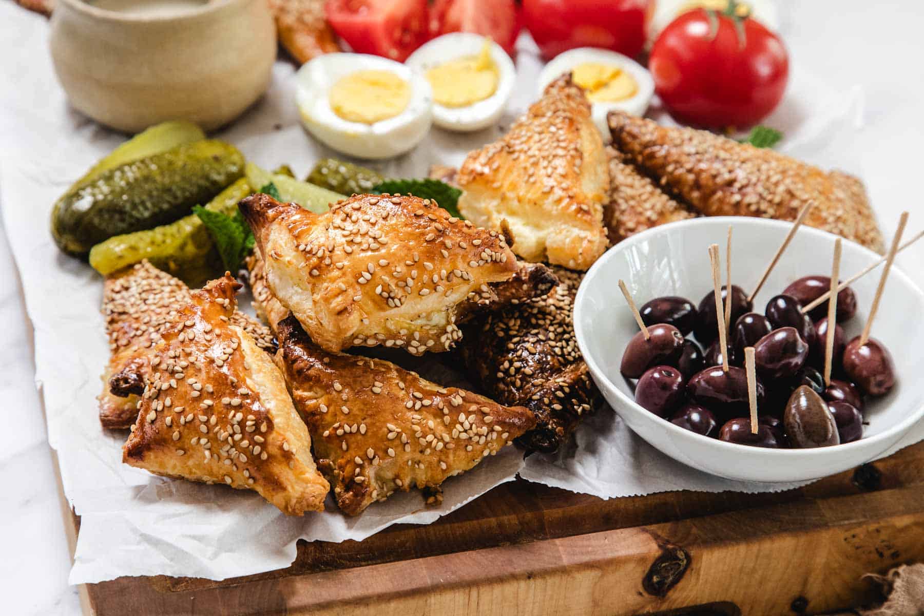 A platter with Bourekas Pinukim—sesame-covered pastry triangles—sliced pickles, black olives in a bowl with toothpicks, hard-boiled egg halves, and fresh tomato wedges on parchment paper.