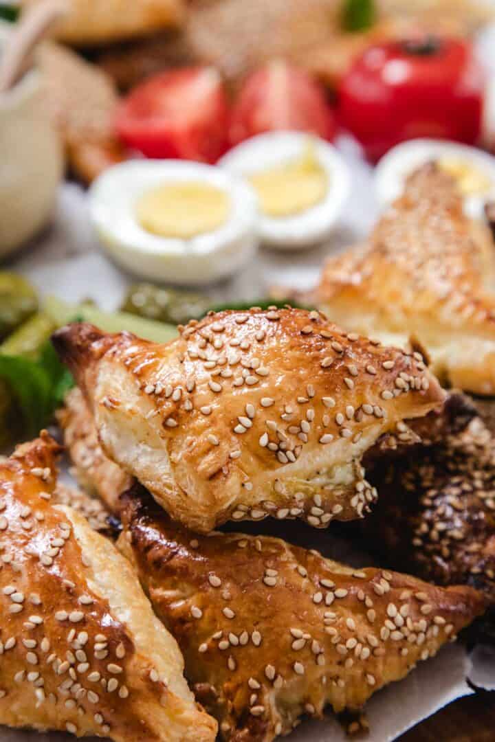 A close-up of golden, triangular Bourekas turnovers topped with sesame seeds. In the blurred background, there are sliced hard-boiled eggs, strawberries, and other assorted foods.