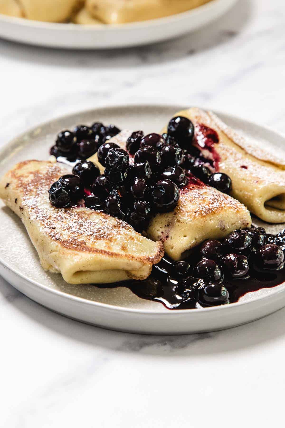 Three cheese blintzes are served on a gray plate, topped with a blueberry compote and a light dusting of powdered sugar. The plate sits on a white marble surface.
