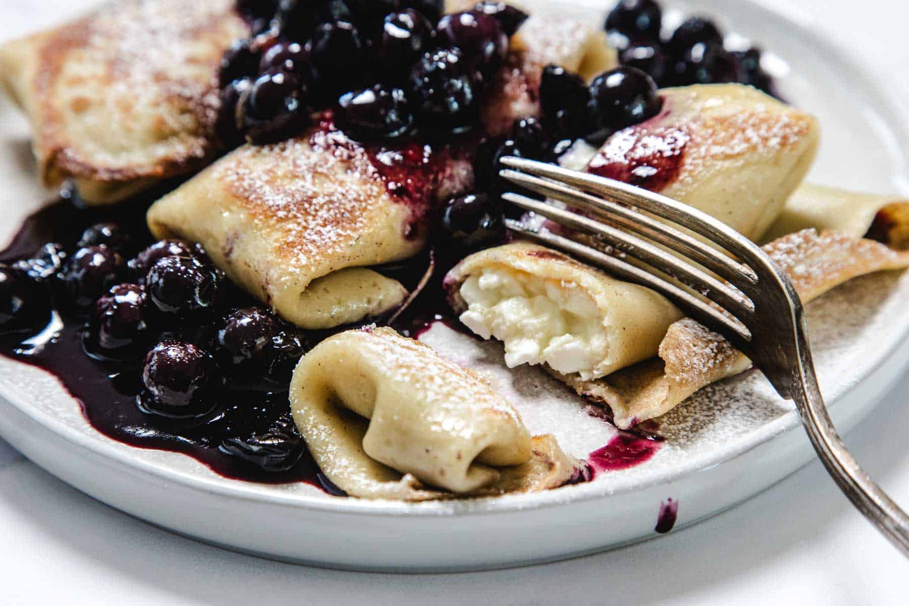 A plate with cheese-filled crepes, or Cheese Blintzes, topped with blueberry sauce and powdered sugar. A fork rests on the plate, with one blintz cut open to reveal the creamy filling inside.