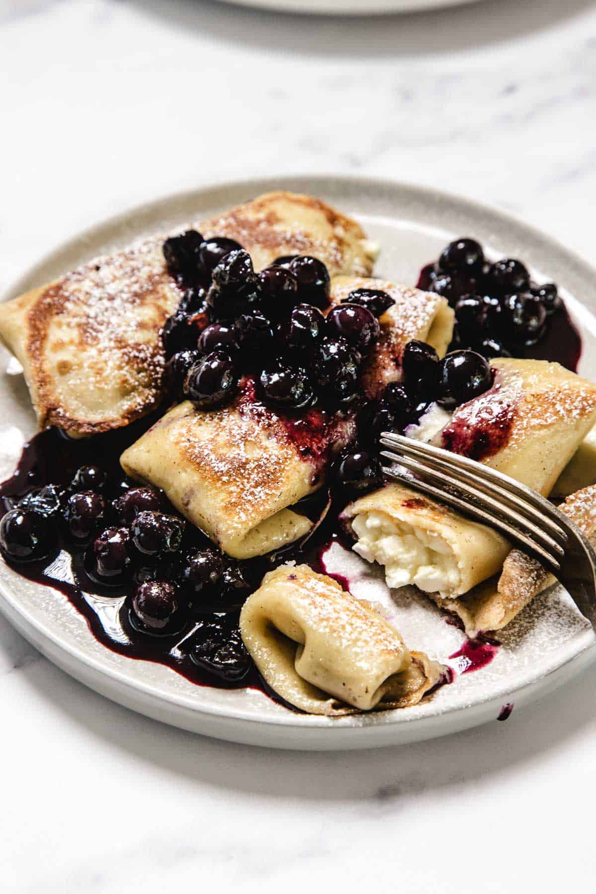 A plate with several cheese-filled crepes, or Cheese Blintzes, topped with cooked blueberries and blueberry sauce, lightly dusted with powdered sugar. A fork rests on the plate, cutting into one of the blintzes.