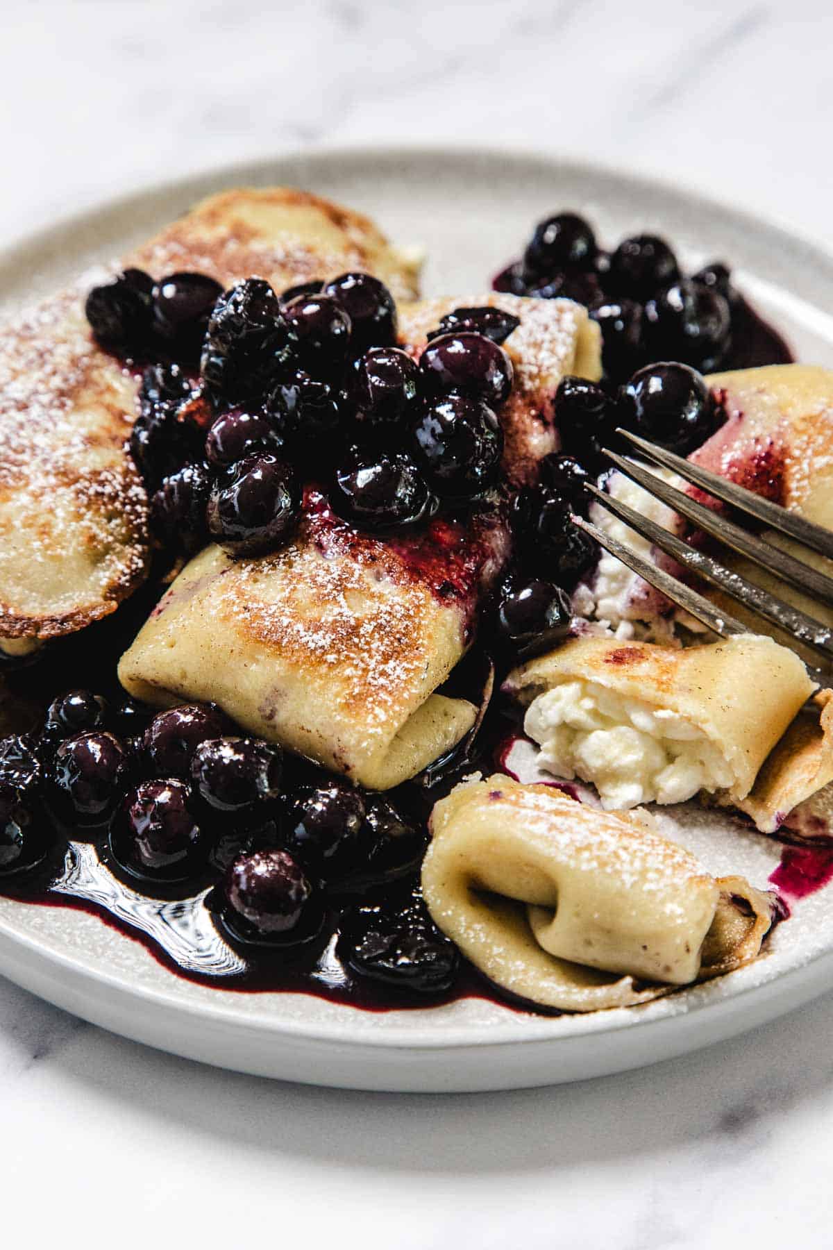 A plate of cheese blintzes filled with creamy cheese, topped with blueberries and powdered sugar, with a fork cutting into one. Blueberry sauce is pooled on the plate.