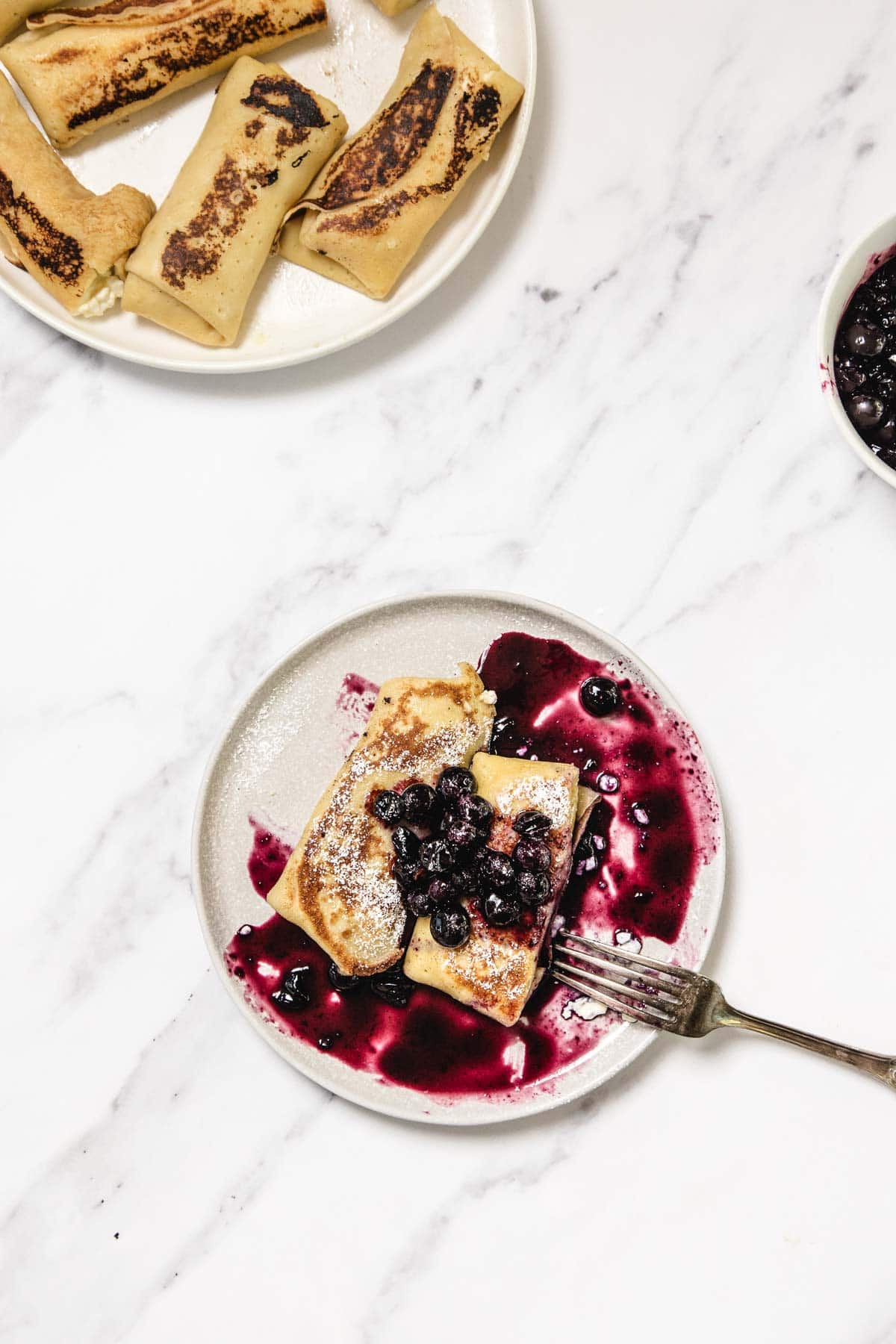 A small plate with two pieces of cheese blintzes topped with blueberry sauce. A fork rests on the plate. In the background, a bowl with more crepes and a bowl of blueberry sauce sit on a white marble surface.