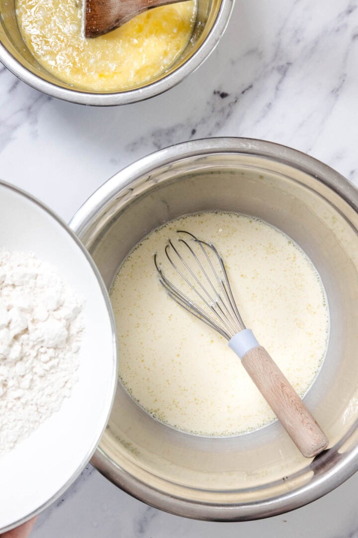A mixing bowl with a whisk in a pale yellow liquid for Cheese Blintzes, a second bowl with a yellow mixture and wooden spoon, and a third bowl holding flour above the first bowl, all arranged on a marble surface.