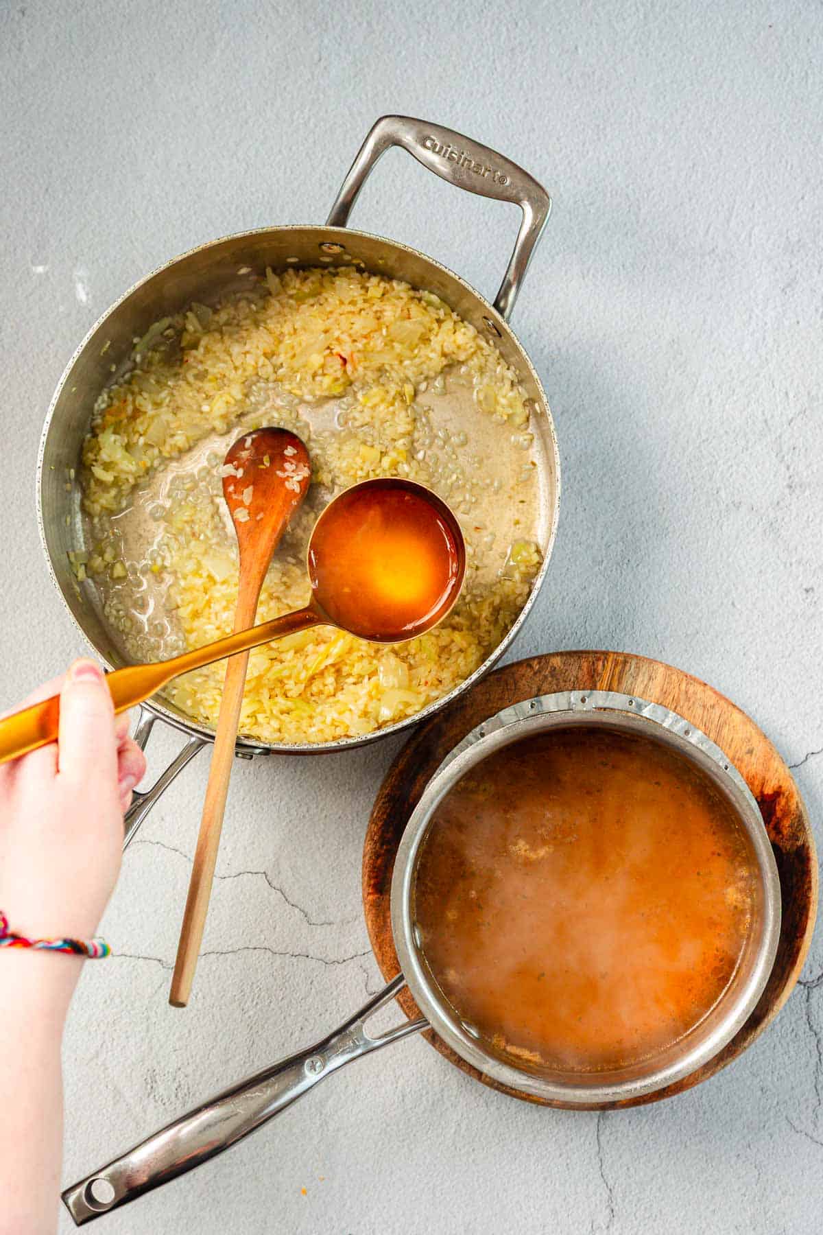 A hand holds a ladle with broth above a saucepan filled with cooked onions and garlic, while another pot with broth sits on a wooden trivet nearby on a gray surface.