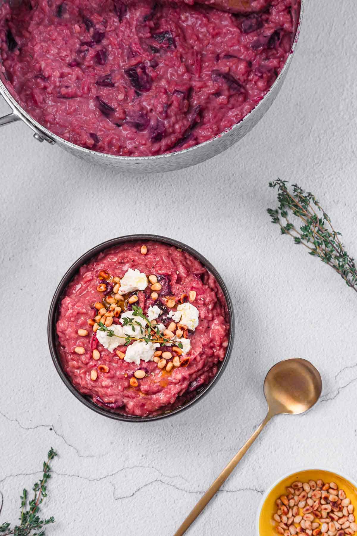 A bowl of purple risotto topped with crumbled cheese, pine nuts, and herbs sits beside a gold spoon. A pot of risotto and a small yellow bowl of pine nuts are nearby, with thyme sprigs on the white surface.