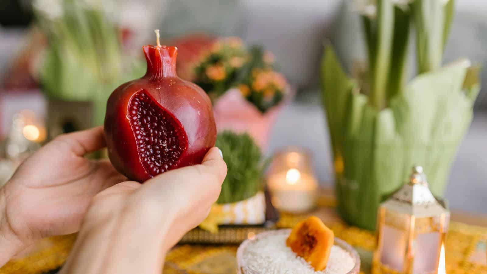 Two hands hold a pomegranate-shaped candle with a carved section showing seeds. In the background, there are flowers, a candle, a lantern, and a glass bowl with rice and a dried orange slice.