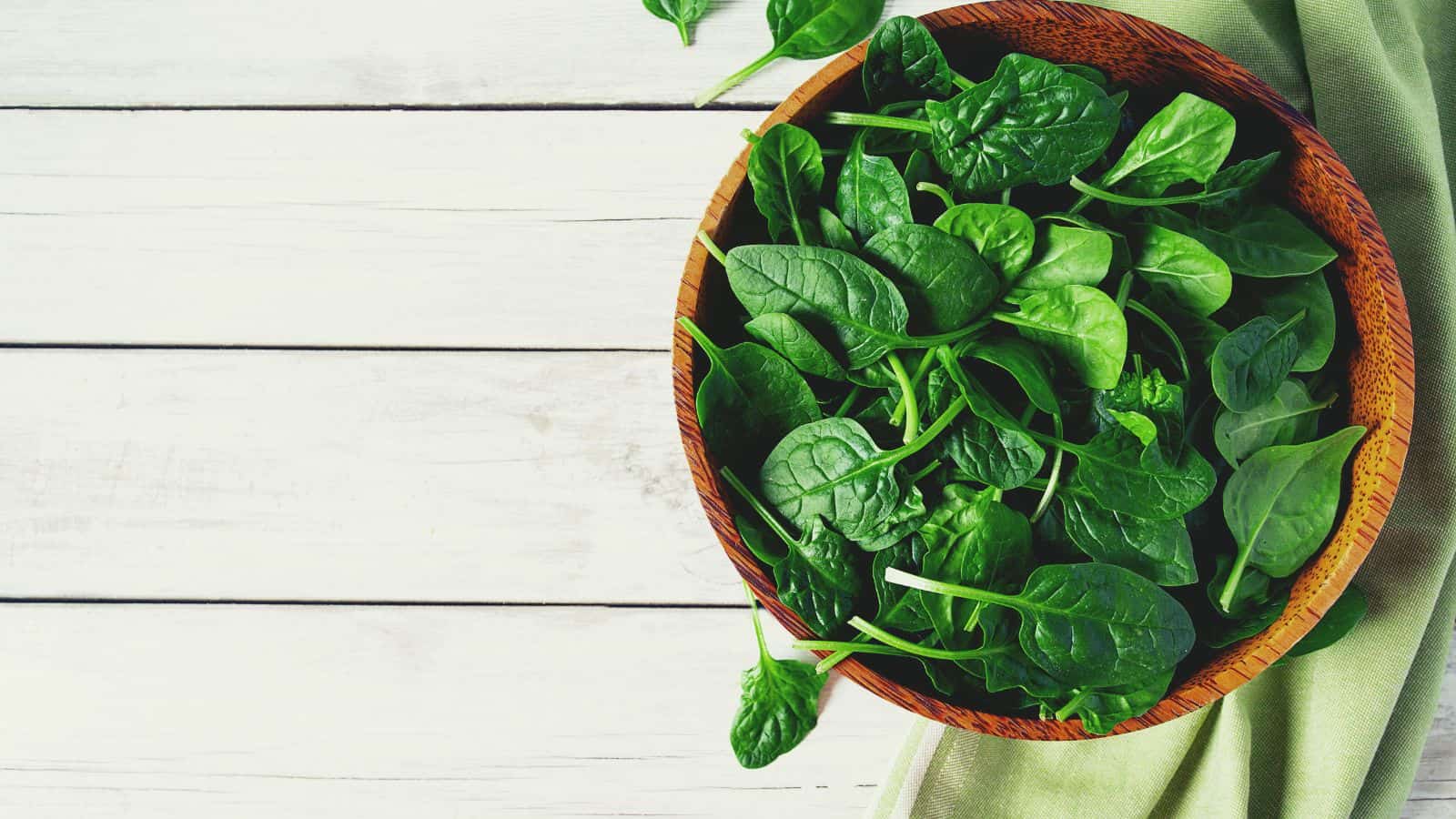A wooden bowl filled with fresh spinach leaves sits on a green cloth atop a white wooden surface.